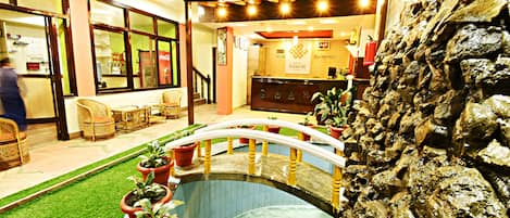 A hotel lobby with a reception desk, a small indoor pool, and several potted plants.