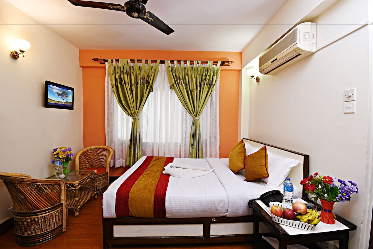 A hotel room with a bed, a desk with a chair, a small table with a vase of flowers, and a wall-mounted air conditioner.