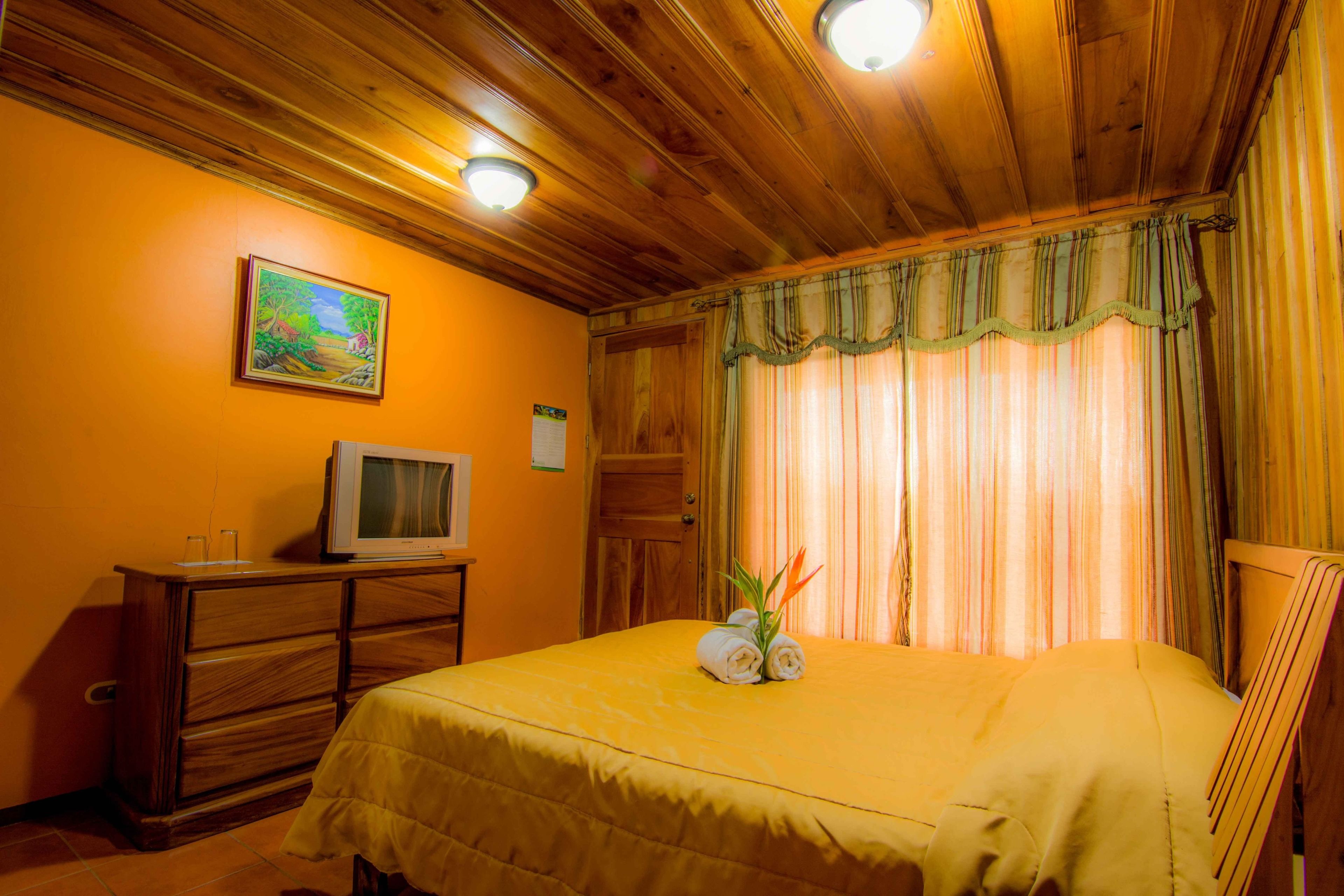 Une chambre d’hôtel avec un plafond en bois, un lit recouvert d’une courtepointe jaune, un téléviseur posé sur une commode en bois et un tableau accroché au mur.