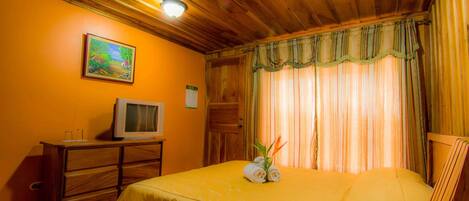 Une chambre d’hôtel avec un plafond en bois, un lit recouvert d’une courtepointe jaune, un téléviseur posé sur une commode en bois et un tableau accroché au mur.