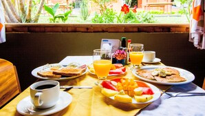 Una mesa de desayuno con tostadas, huevos, panqueques y fruta, junto con tazas de café y jugo de naranja.