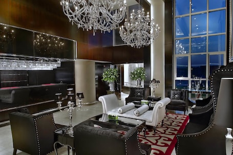 A luxurious living room with a large flat-screen TV, elegant chandeliers, and a large window offering a city view.