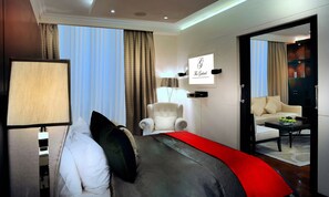 A hotel room with a bed, a chair, a desk with a lamp, and a view of another room.