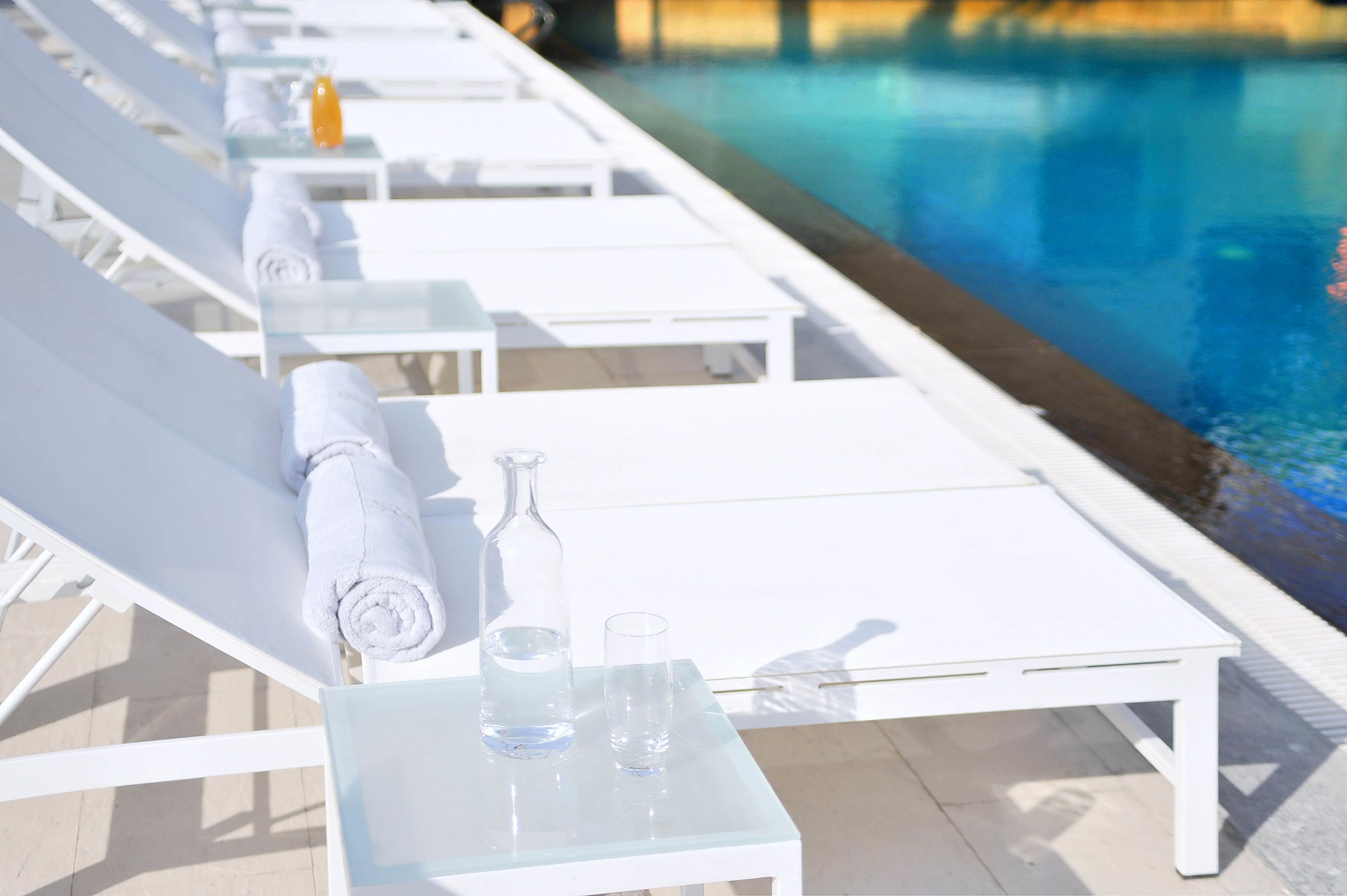 A row of white sun loungers with towels and a glass bottle and glass on a table by a pool.