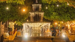 Un décor extérieur nocturne avec une fontaine, des guirlandes lumineuses et des espaces aménagés pour s’asseoir.