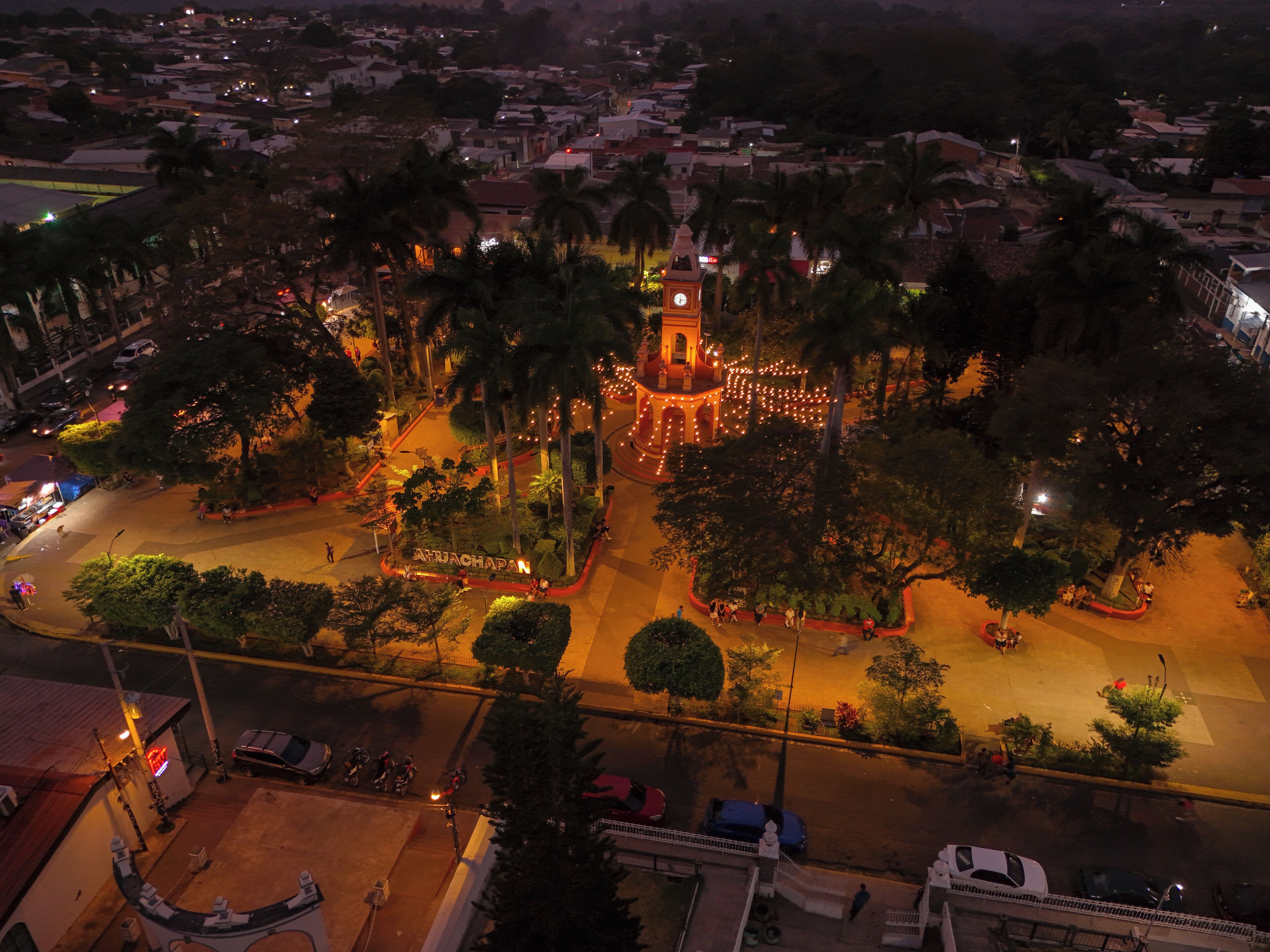 A nighttime aerial view of a town with a prominent clock tower, surrounded by palm trees and illuminated streets.