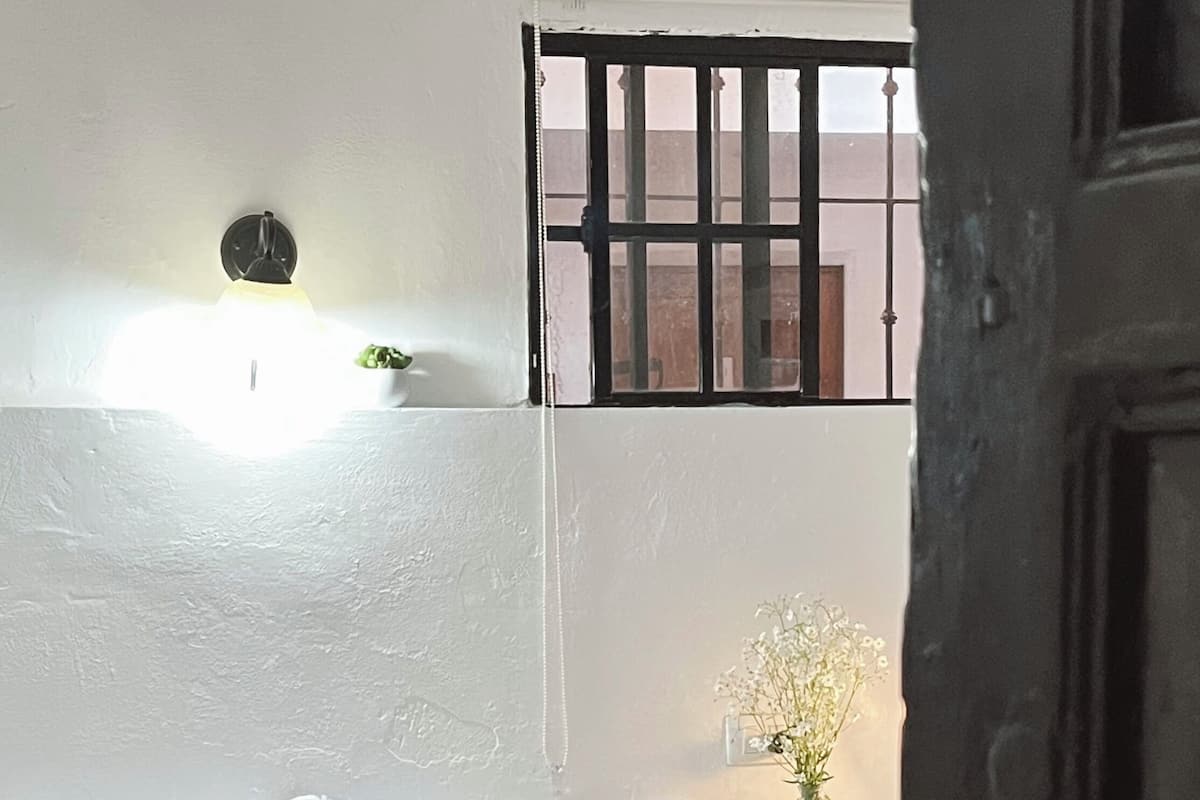 A bedroom with a bed, a nightstand, a wall-mounted light, a window with bars, and a door.