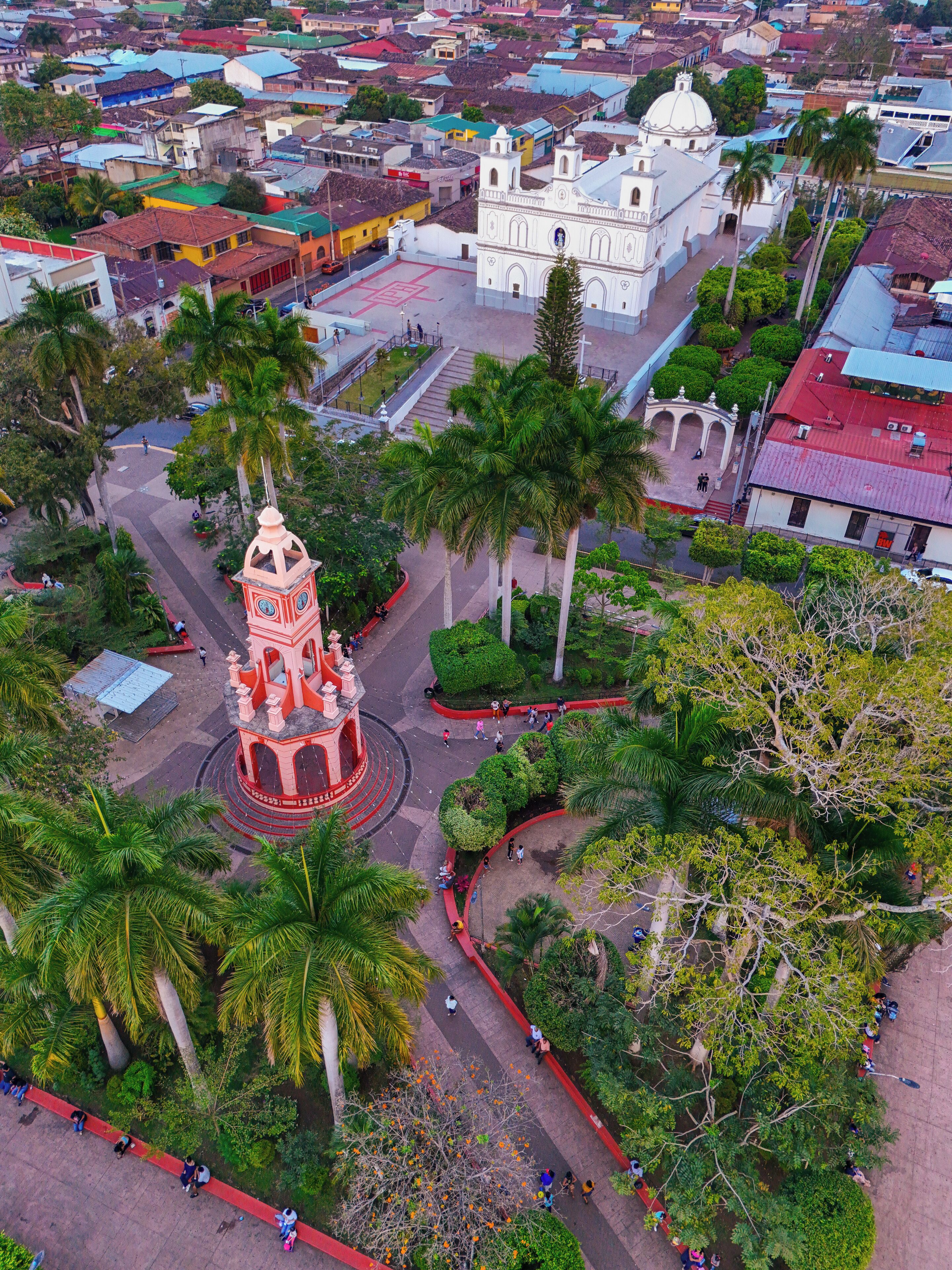 An aerial view of a cityscape with a prominent white church with a dome, a central red and white tower, and surrounding greenery.