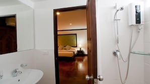 A hotel bathroom with a sink, a shower, and a door leading to a bedroom with a bed and nightstand. - Cucumber Inn (Pattaya)