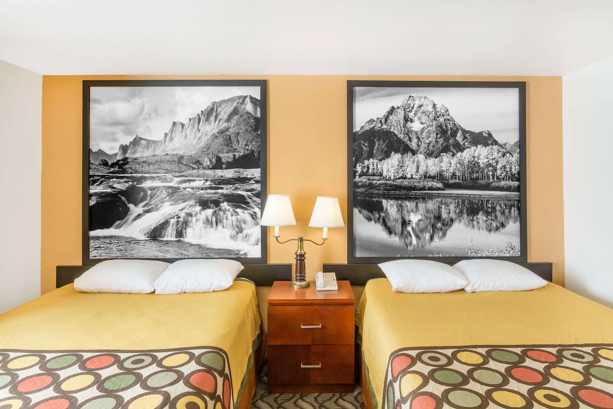 A hotel room with two beds, a nightstand with a lamp, and two large framed pictures of mountains and a lake.