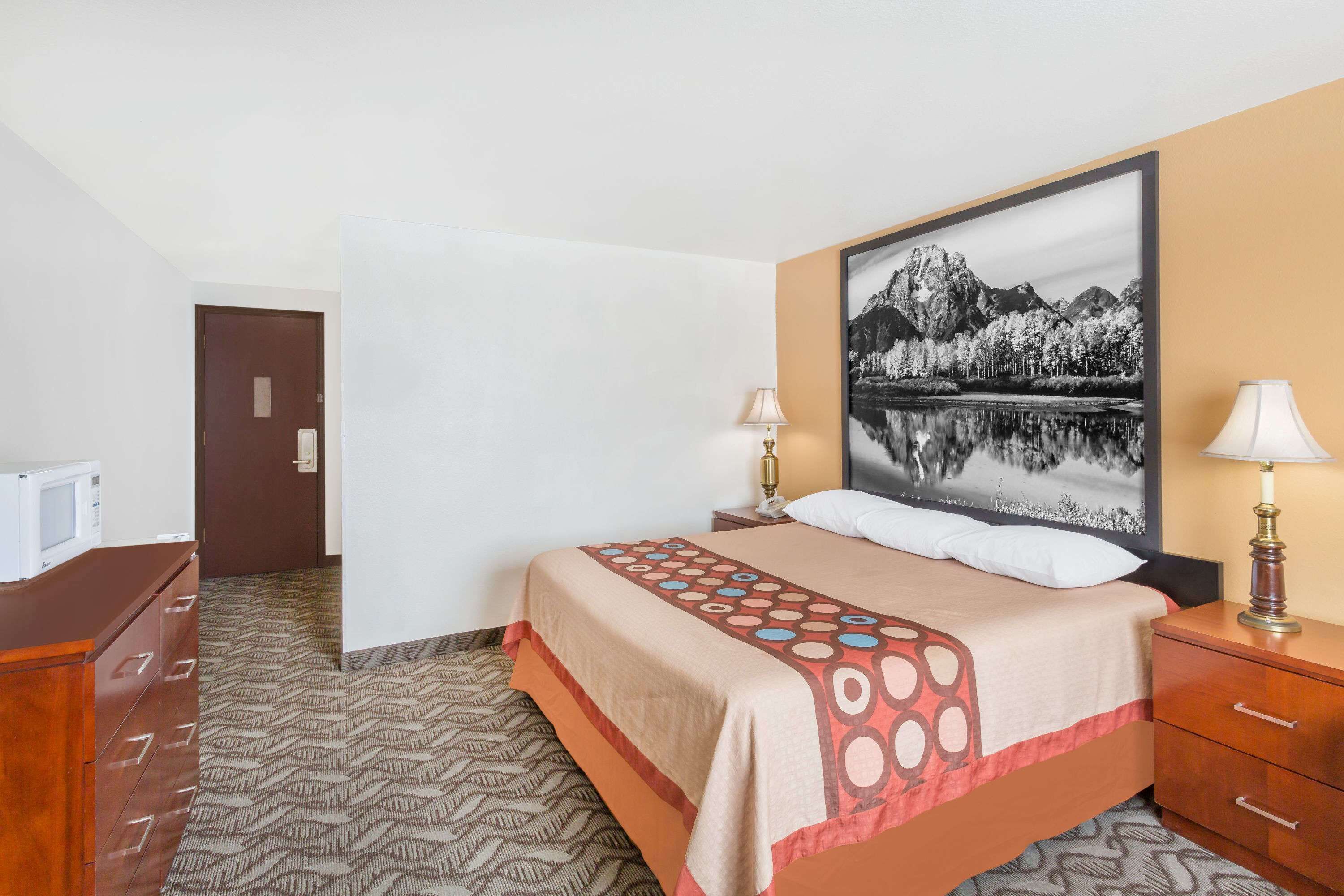 A hotel room with a bed, two bedside lamps, a wooden nightstand, a dresser, and a large framed picture on the wall.