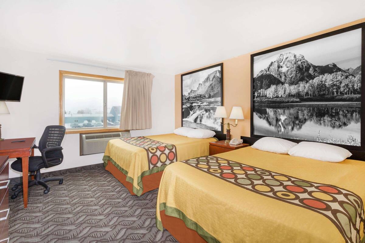 A hotel room with two beds, a desk, a chair, a TV, and large framed pictures of mountains and a lake.