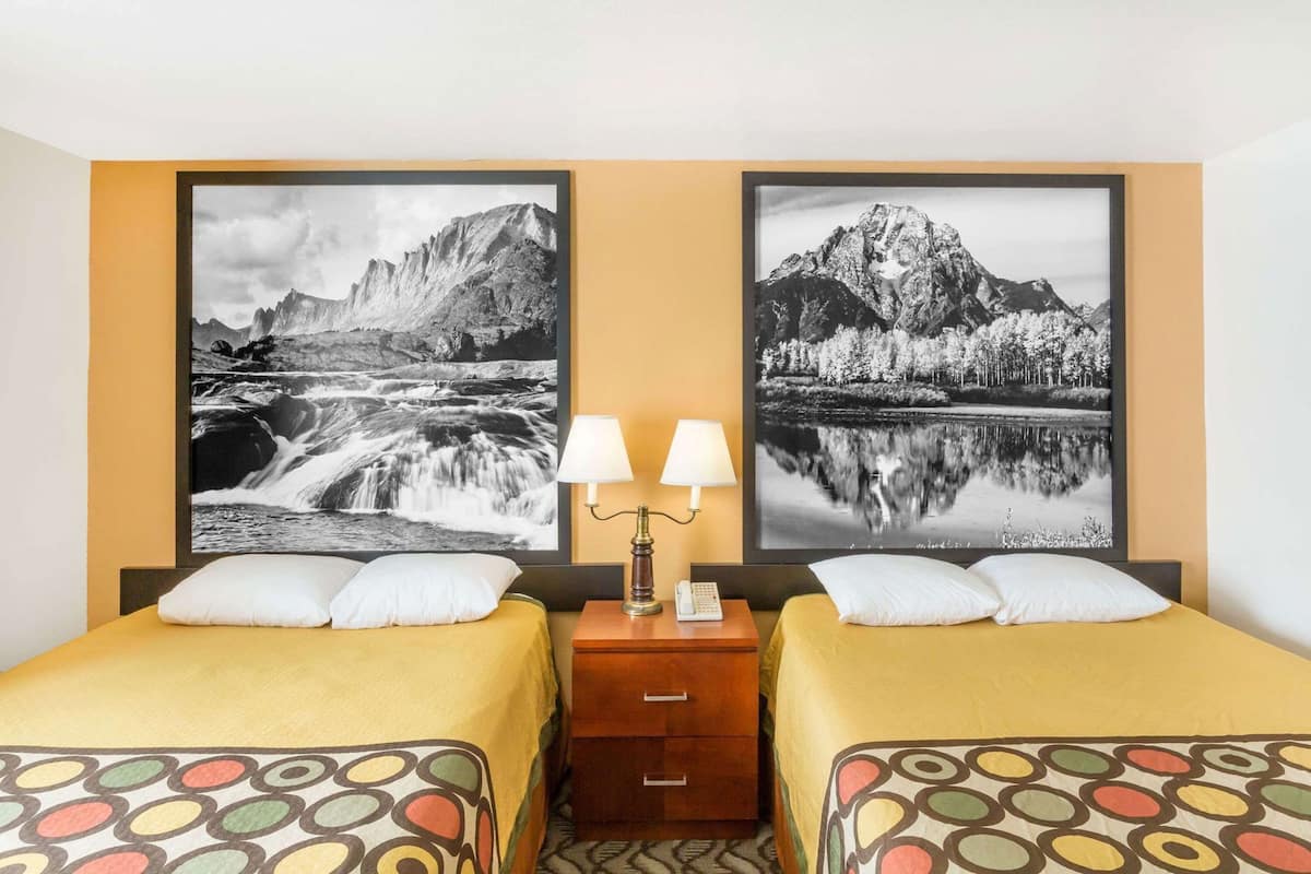 A hotel room with two beds, a nightstand with a lamp, and two large landscape paintings on the wall.