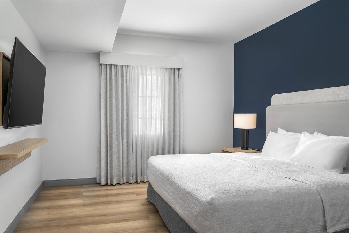 A hotel room with a bed, a TV mounted on the wall, a bedside table with a lamp, and a window with curtains.