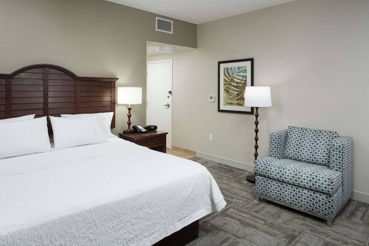 A hotel room with a large bed, bedside lamps, a chair, a door, and a framed artwork on the wall.