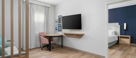 Quarto de hotel com escrivaninha, cadeira, cama, televisão e mesa de cabeceira.