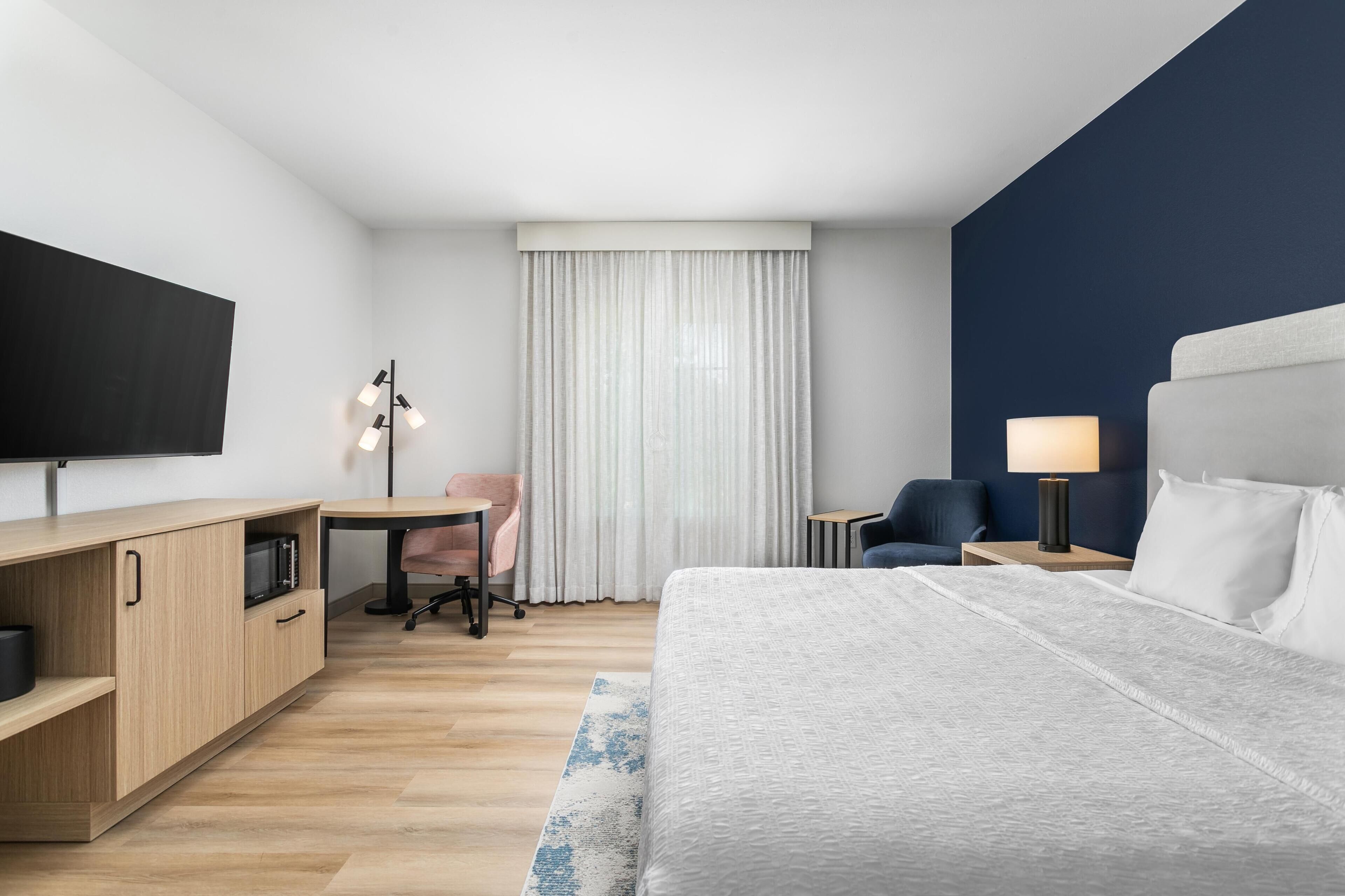 Habitación de hotel con cama, escritorio, silla, televisión, lámpara y ventana con cortinas.