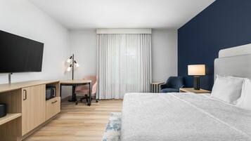Habitación de hotel con cama, escritorio, silla, televisión, lámpara y ventana con cortinas.