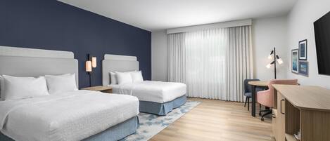 A hotel room with two beds, a desk, a chair, a lamp, and a window with curtains.