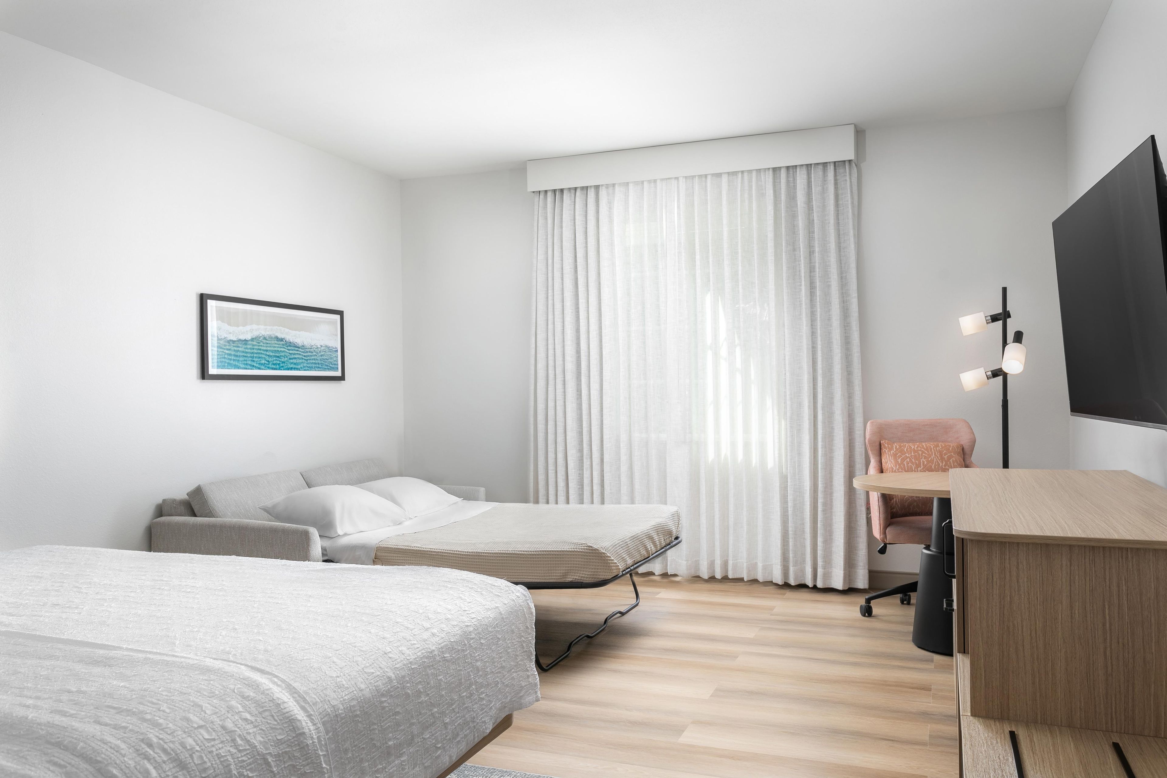 Ein modernes Hotelzimmer mit Bett, Schreibtisch, Stuhl, Fernseher und einem fensterbepflanzten Fenster.