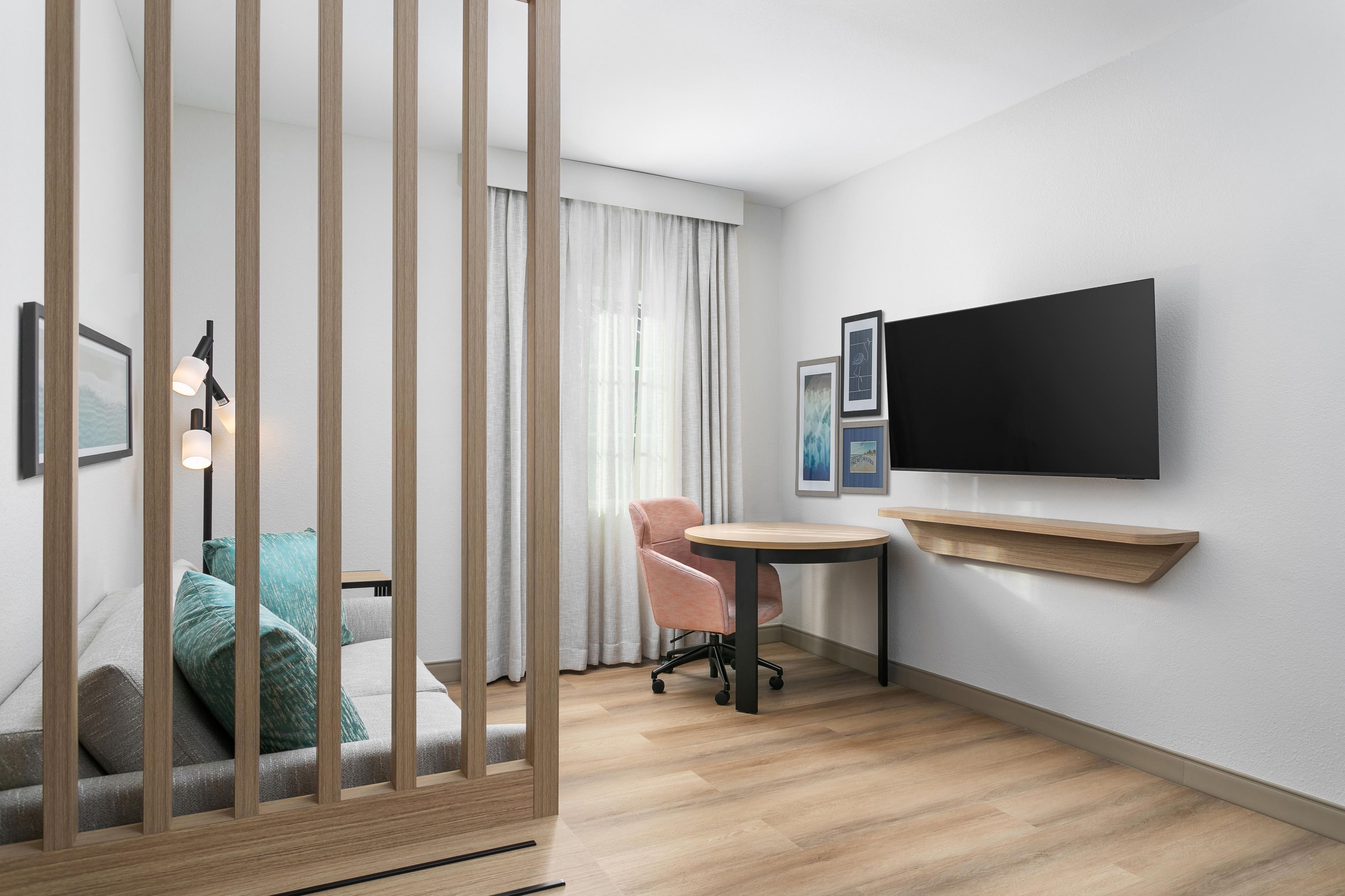 Una habitación de hotel moderna con piso de madera, un escritorio con silla, televisor montado en la pared y una ventana con cortinas.