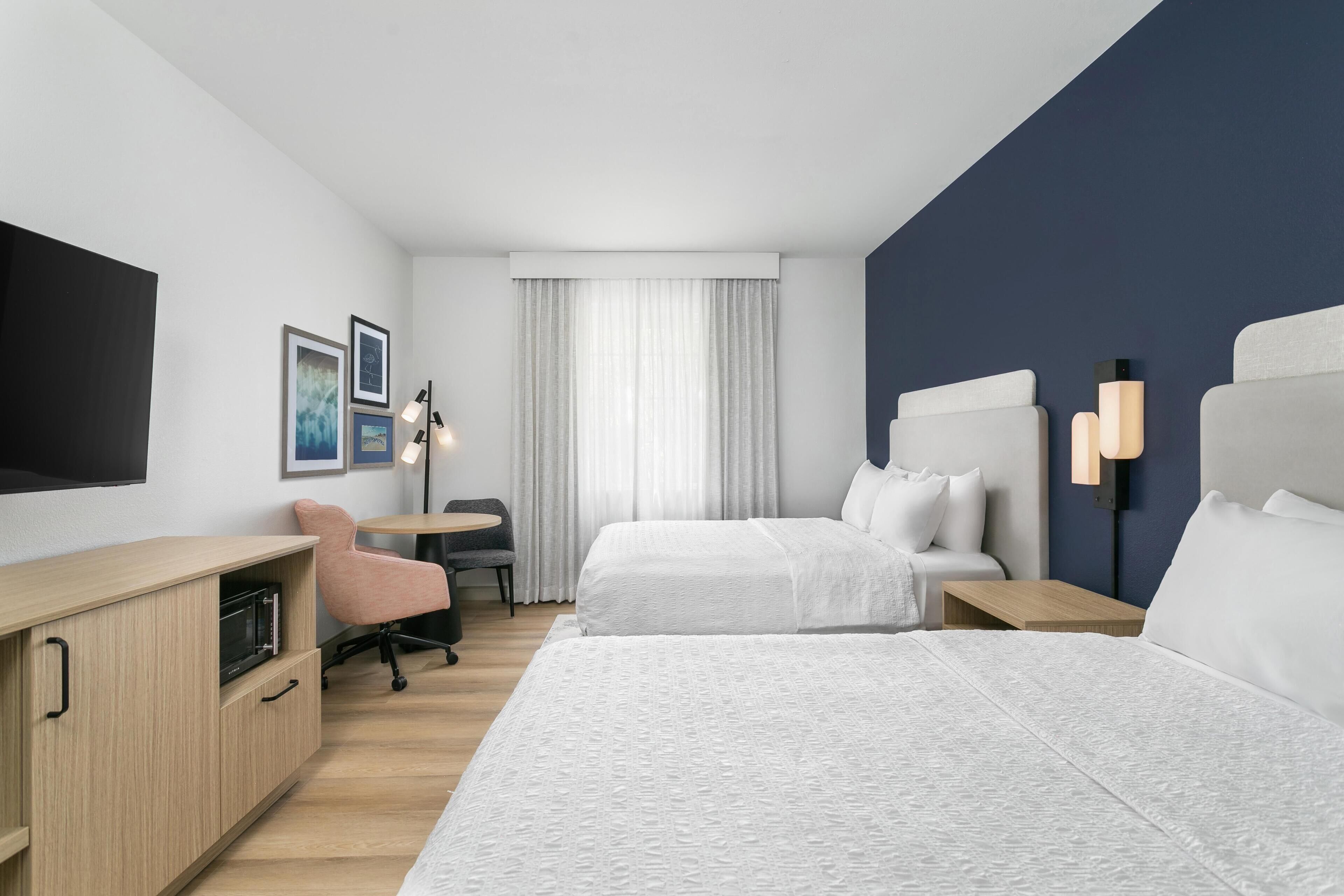 Une chambre d’hôtel avec deux lits, un bureau, une chaise, une télévision et une fenêtre avec des rideaux.