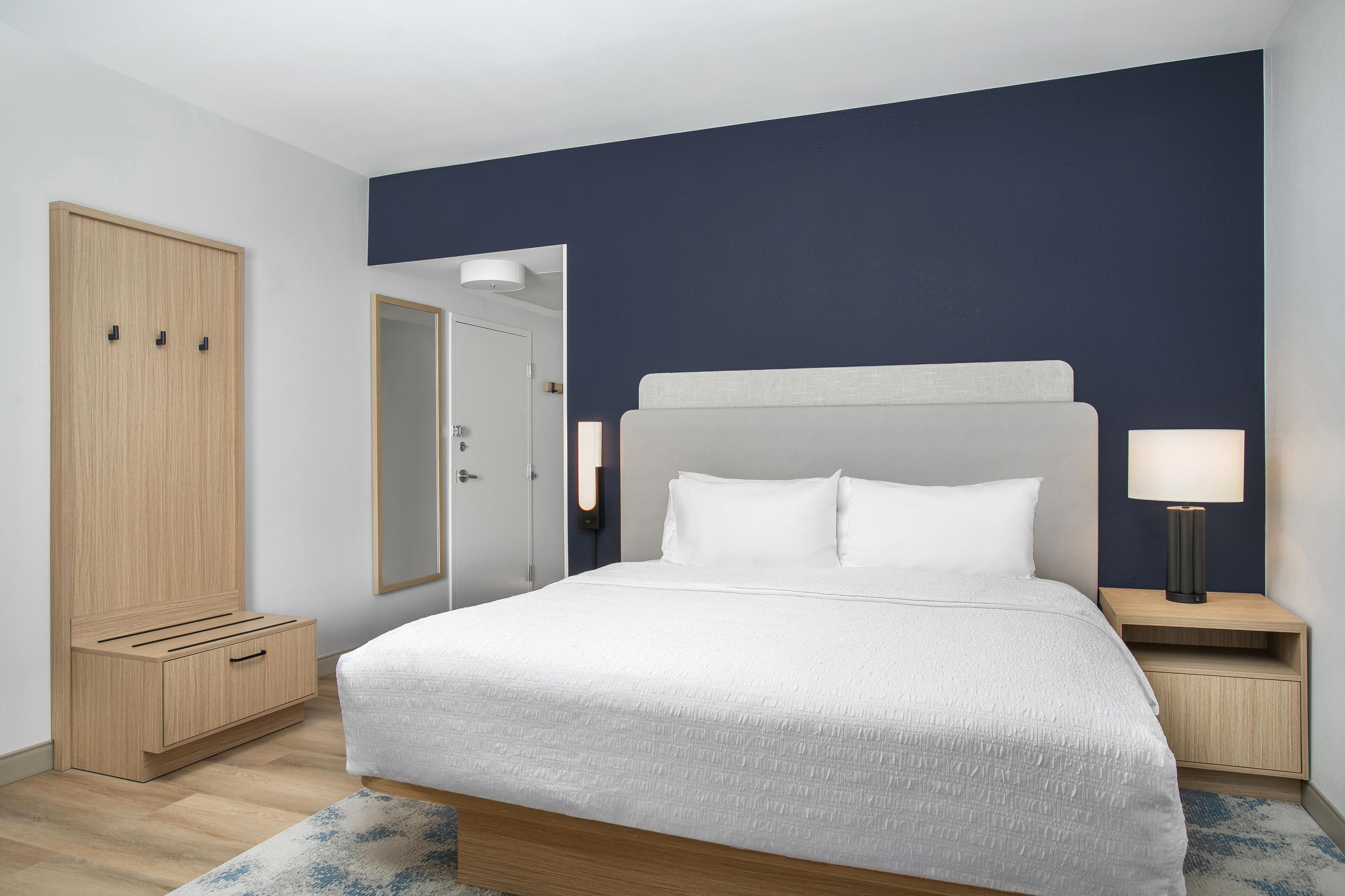 Una habitación de hotel moderna con una cama grande, un armario, un espejo y una mesita de noche con una lámpara.