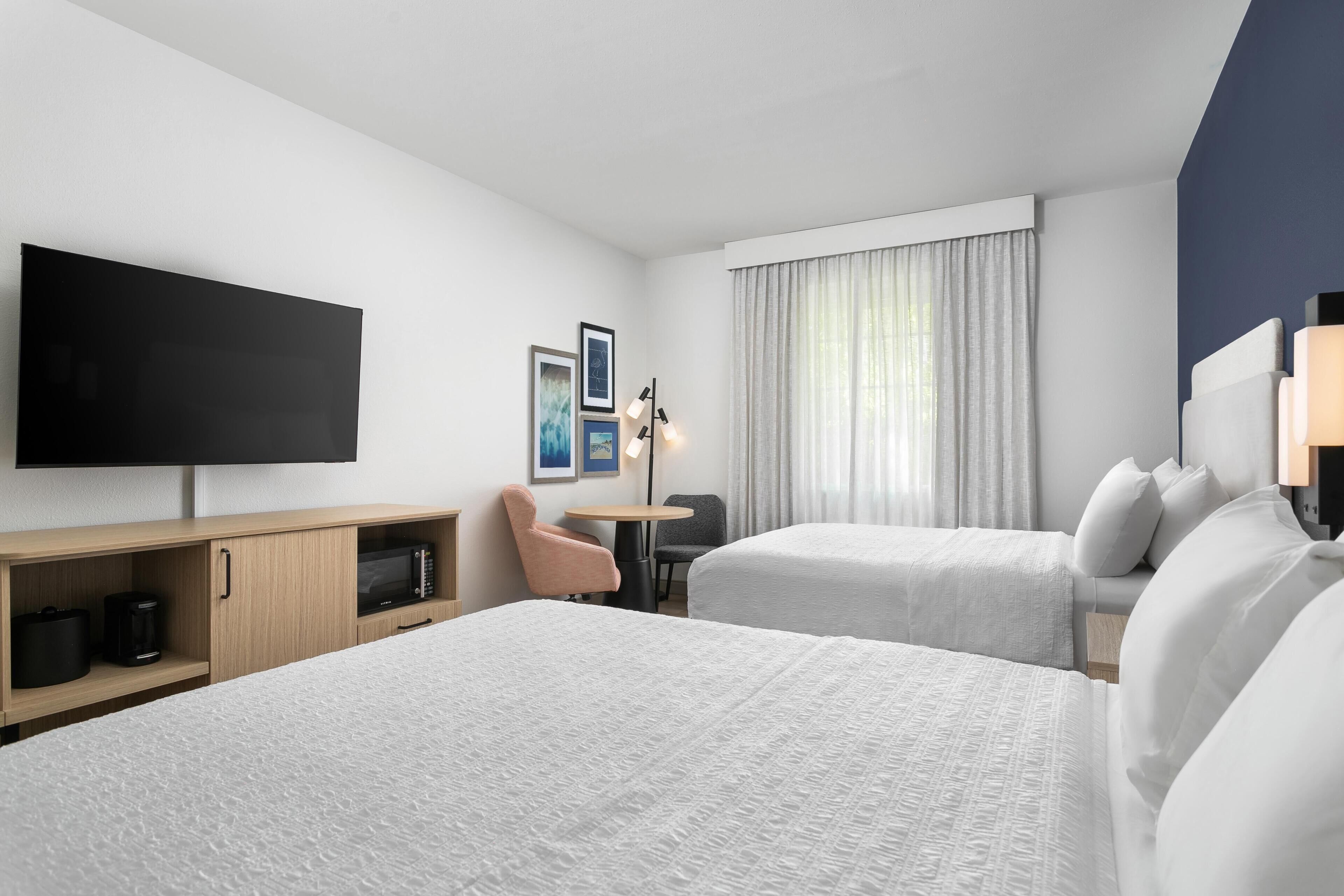 A hotel room with two beds, a flat-screen TV, a desk with a coffee maker, a chair, and a window with curtains.