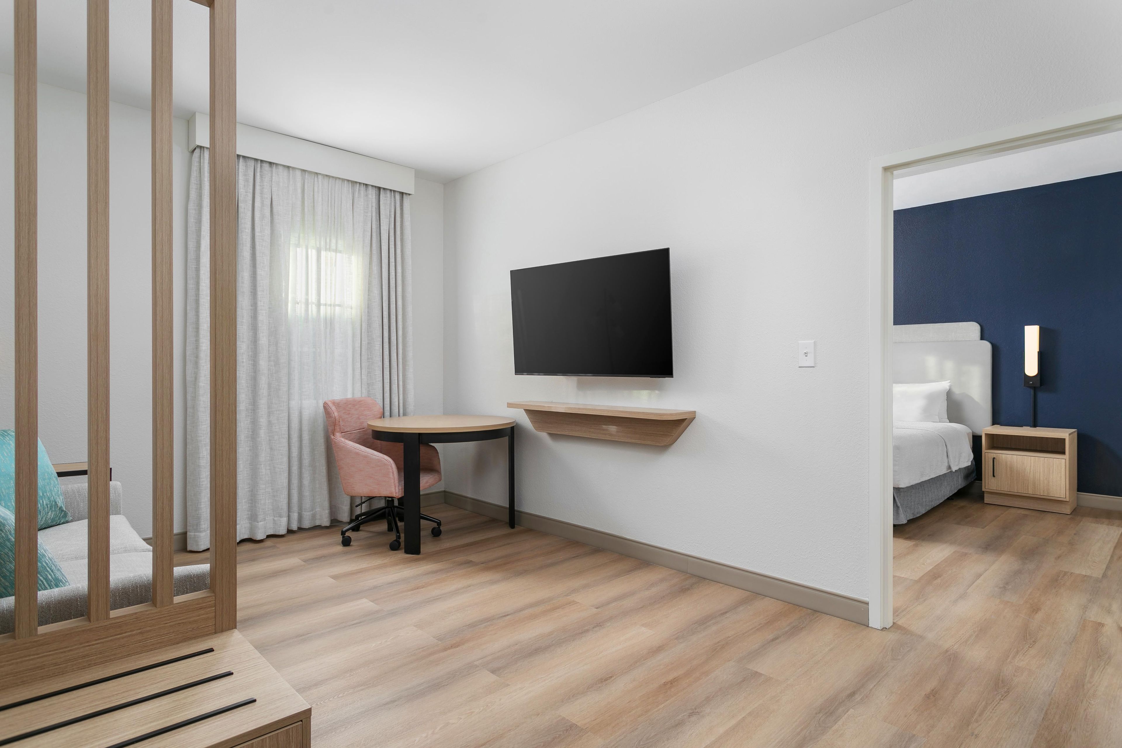 A hotel room with a bed, a desk with a chair, a television mounted on the wall, and a window with curtains.