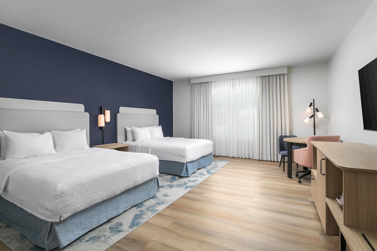 A hotel room with two beds, a desk, a chair, and a large window with curtains.