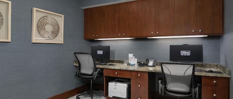 A hotel room with a desk, two chairs, a printer, and framed artwork on the wall.