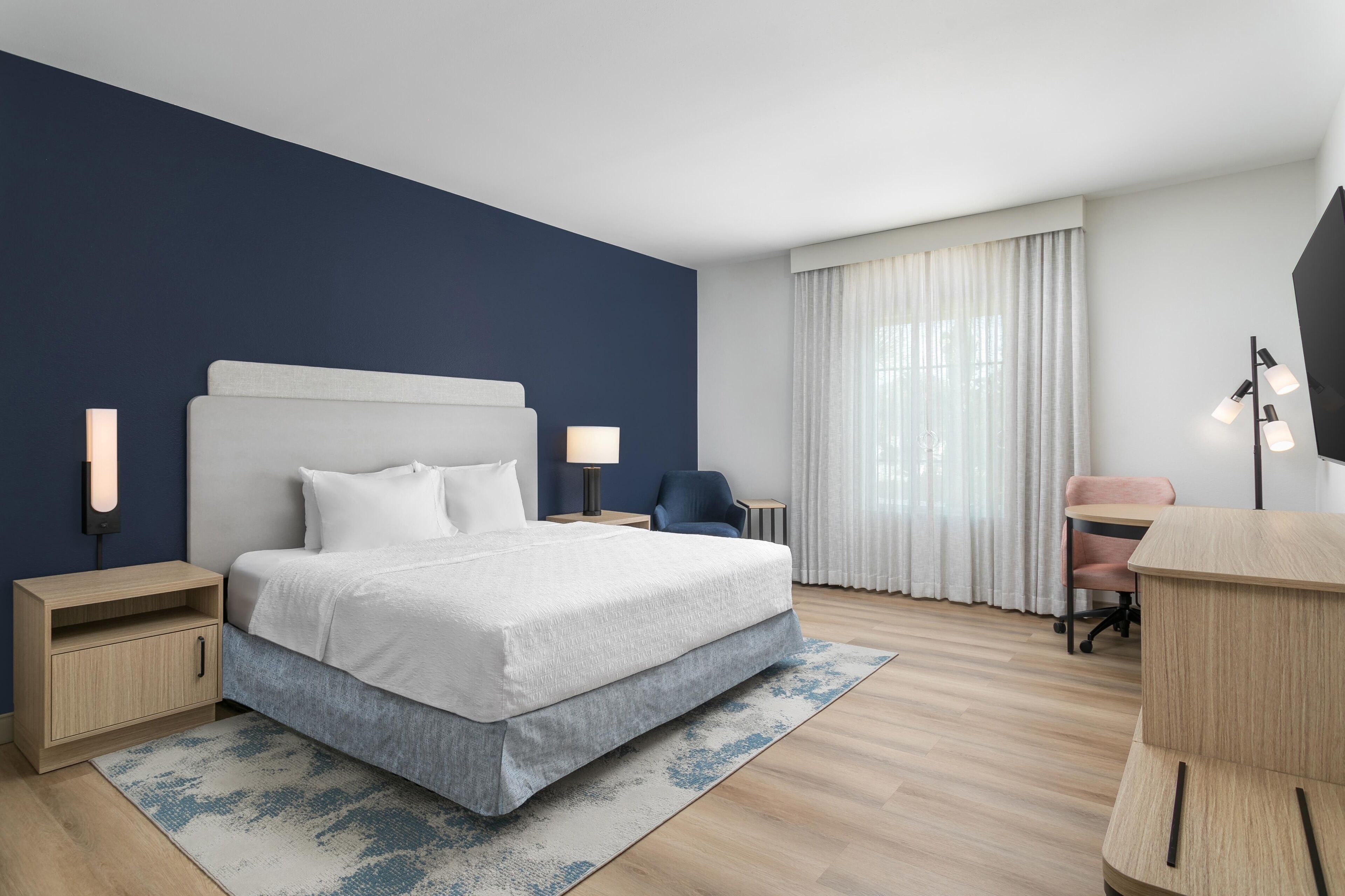 A hotel room with a bed, bedside tables, a desk, a chair, and a large window with curtains.