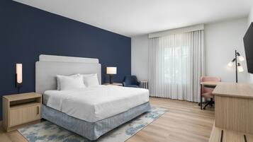 A hotel room with a bed, bedside tables, a desk, a chair, and a large window with curtains.