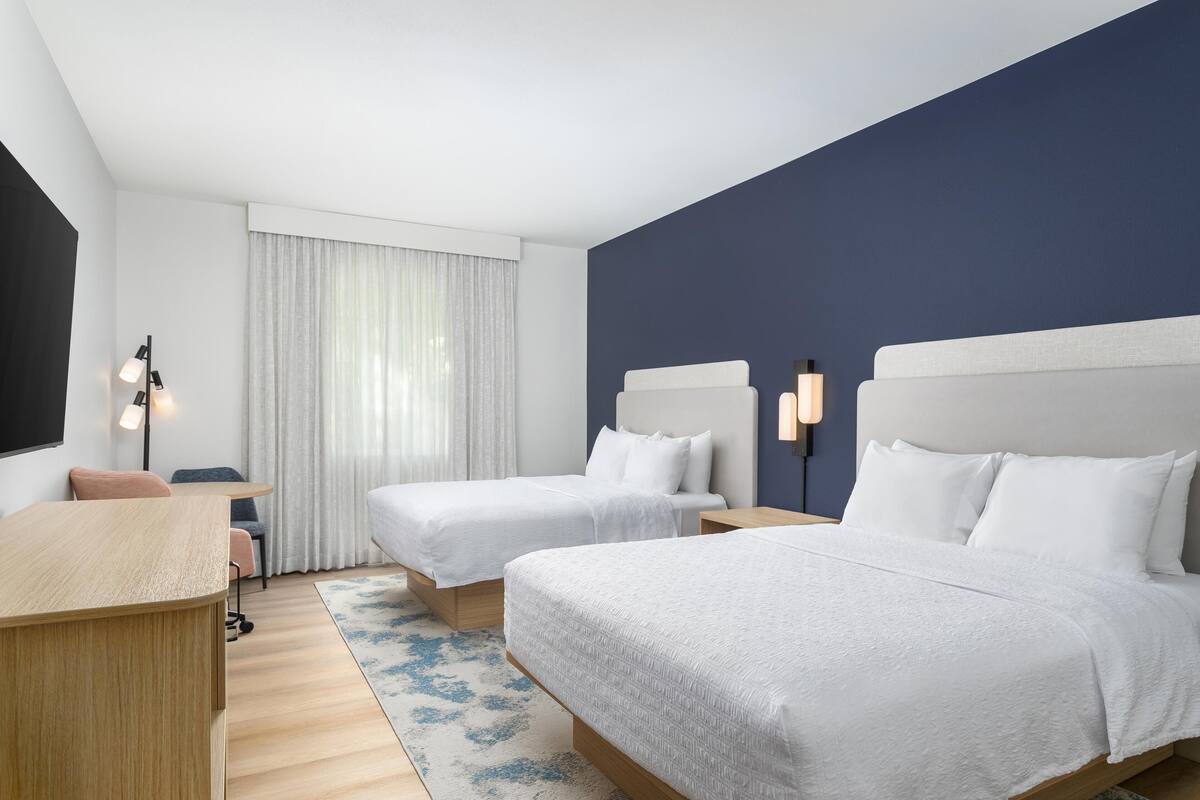 A hotel room with two beds, a desk, a chair, a lamp, and a window with curtains.