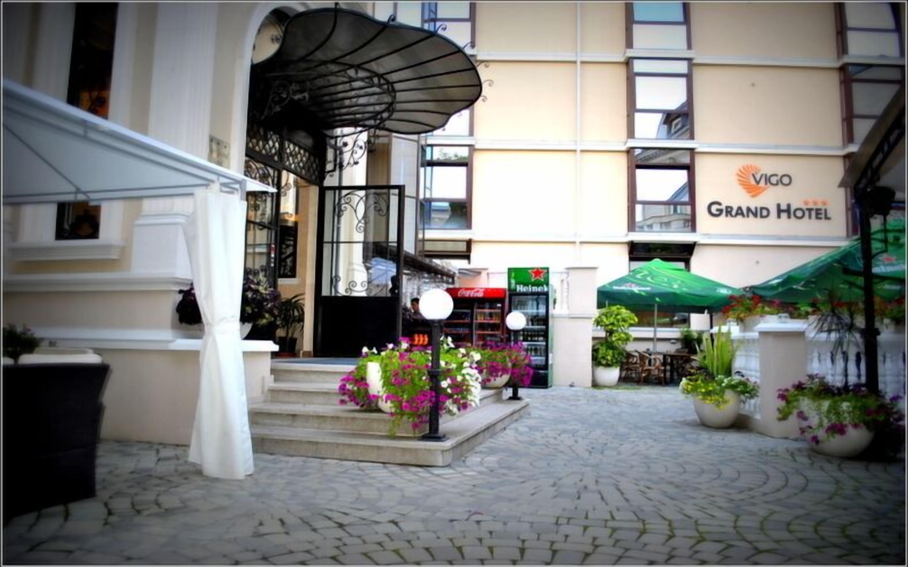 Entrada del Grand Hotel con un patio adoquinado, una escalera cubierta y zonas de estar al aire libre con sombrillas.