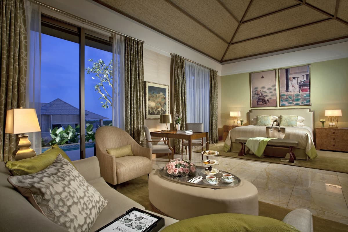 A hotel room with a large window, a bed, a sofa, a dining table with a tray of food, and a painting on the wall.