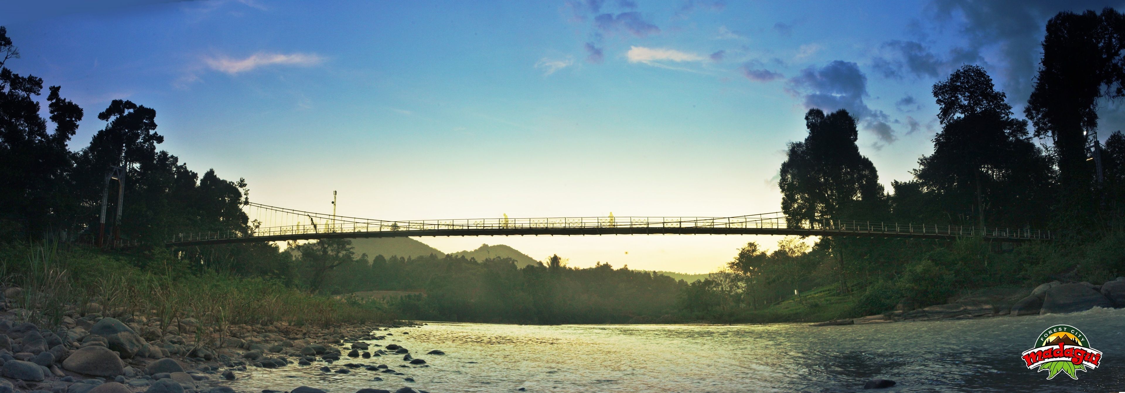 Un puente colgante sobre un río, con árboles aledaños y un logotipo en la esquina.