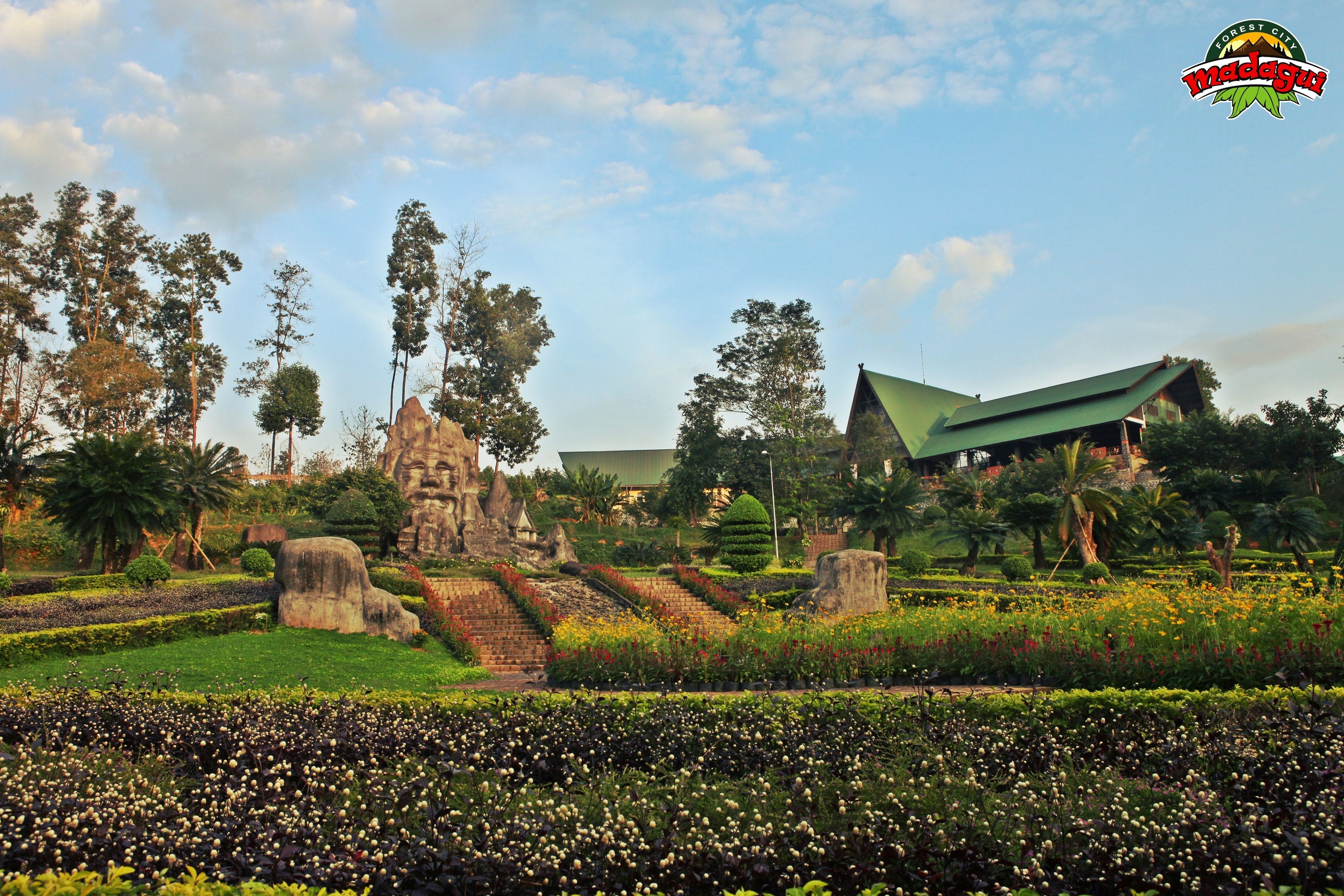Un jardín ajardinado con un monumento de piedra, un edificio con techo verde y una variedad de plantas y flores.