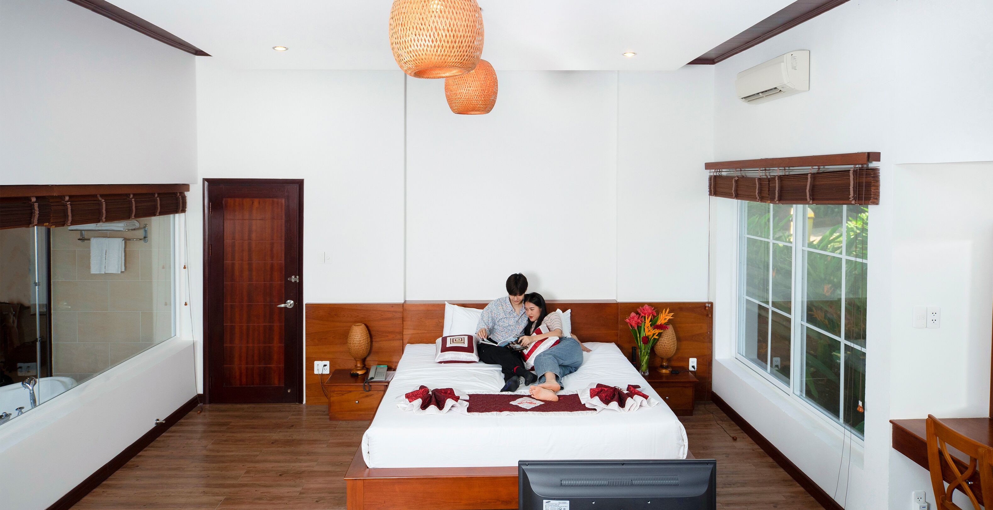 A modern bedroom with a large bed, a wooden headboard, a bedside table with a lamp, and a large window with a view of greenery.
