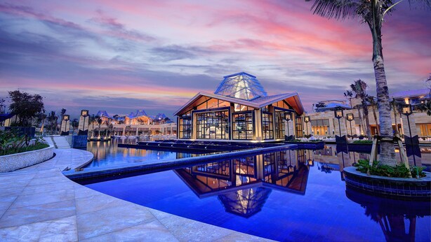 A modern hotel with a large pool, a dome-shaped glass structure, and palm trees. - The Mulia Bali (Nusa Dua)