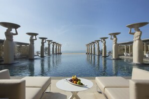 A poolside area with lounge chairs and a table with fruit, surrounded by statues holding umbrellas. - The Mulia Bali (Nusa Dua)