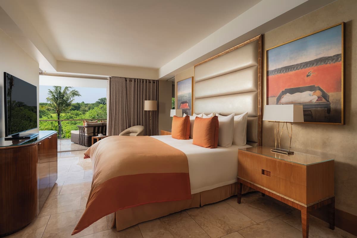 A hotel room with a bed, a desk, a TV, and a view of greenery.