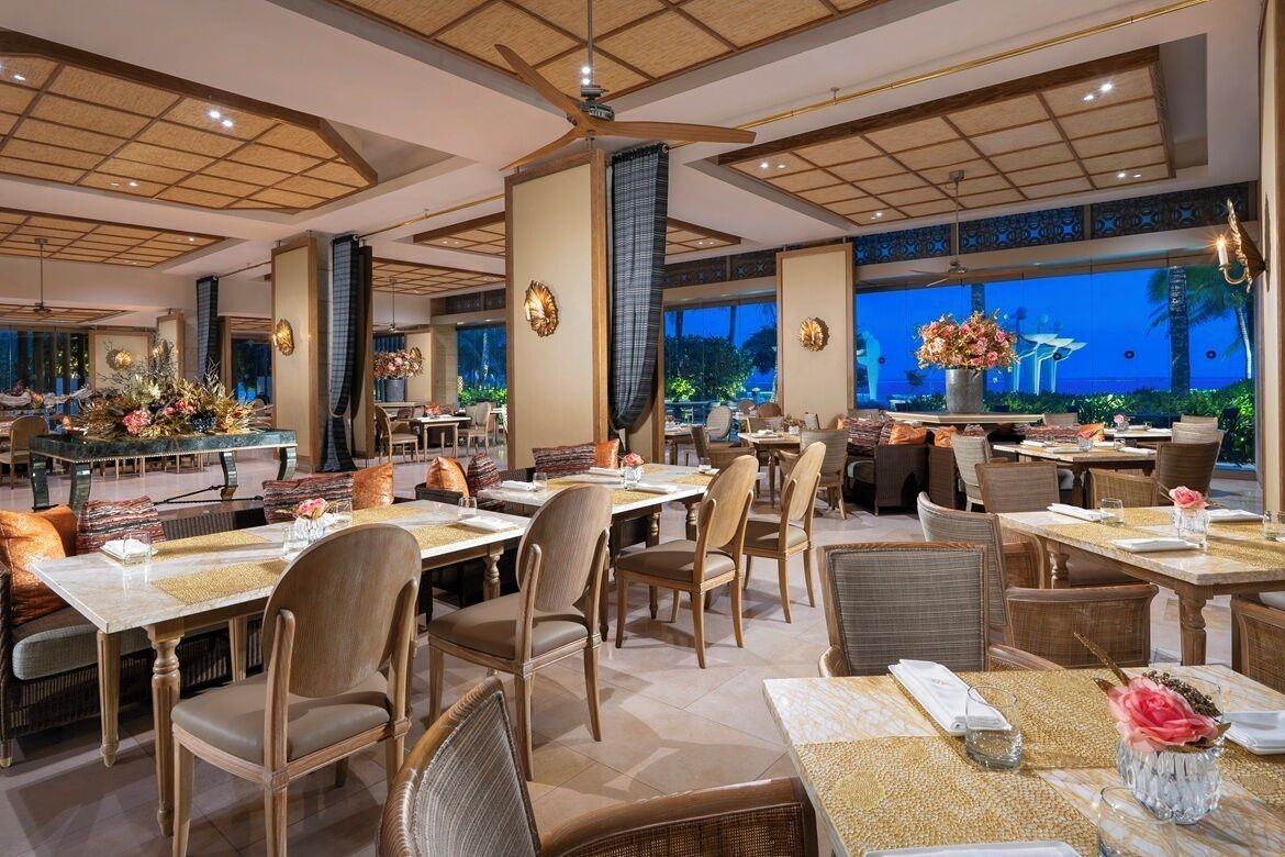 Um restaurante espaçoso com várias mesas e cadeiras, grandes janelas com vista para o exterior e arandelas decorativas nas paredes.