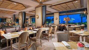 Um restaurante espaçoso com várias mesas e cadeiras, grandes janelas com vista para o exterior e arandelas decorativas nas paredes.