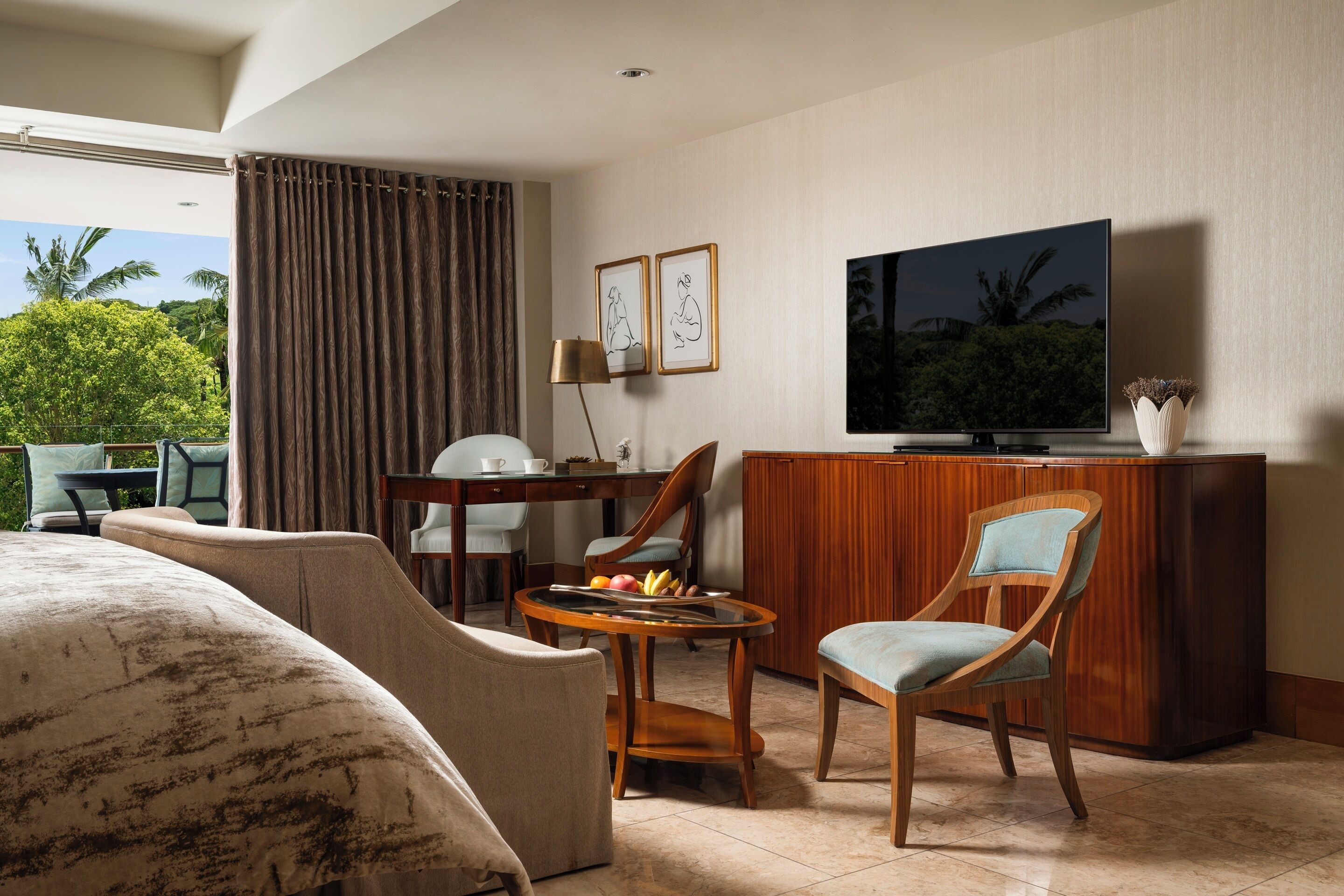 A hotel room with a bed, a TV, a desk with a lamp, a chair, and a view of greenery outside.