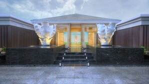 A modern building with a glass facade, a central entrance with steps, and two large white sculptures on either side. - The Mulia Bali (Nusa Dua)