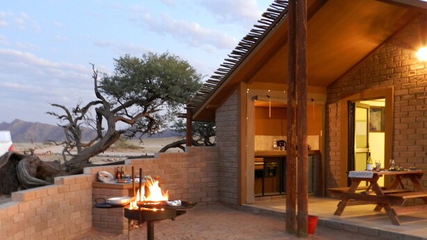 A desert-style outdoor area with a fire pit, a wooden table, and a brick wall. - Desert Camp (Sesriem)