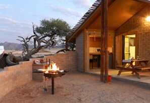Un espacio al aire libre de estilo desértico con un brasero, una mesa de madera y una pared de ladrillo.