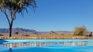 A swimming pool with clear blue water, surrounded by a stone wall, with a view of mountains in the background. - Desert Camp (Sesriem)