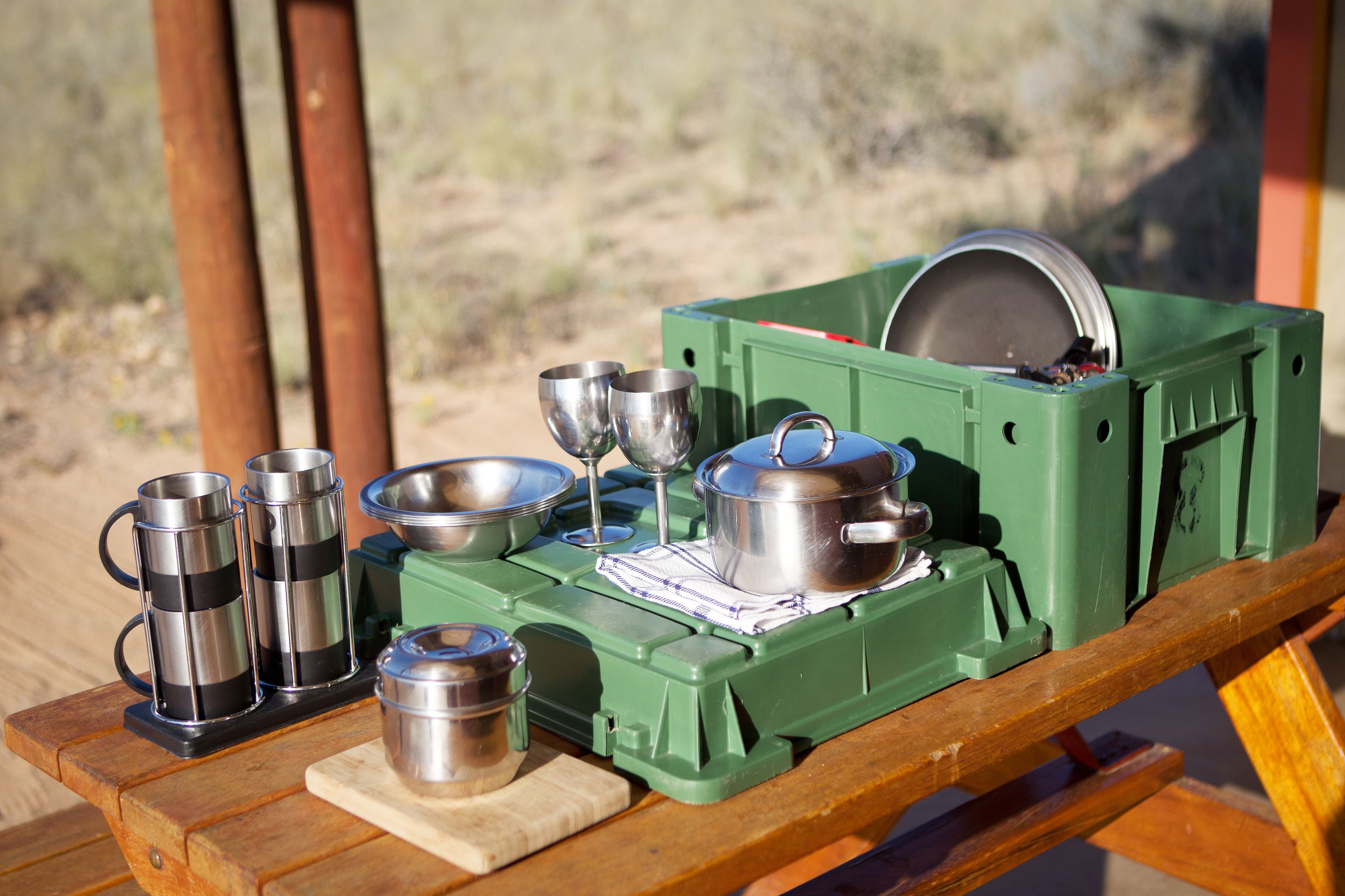 A camping setup with a green portable stove, cooking pots, and stainless steel mugs on a wooden table.